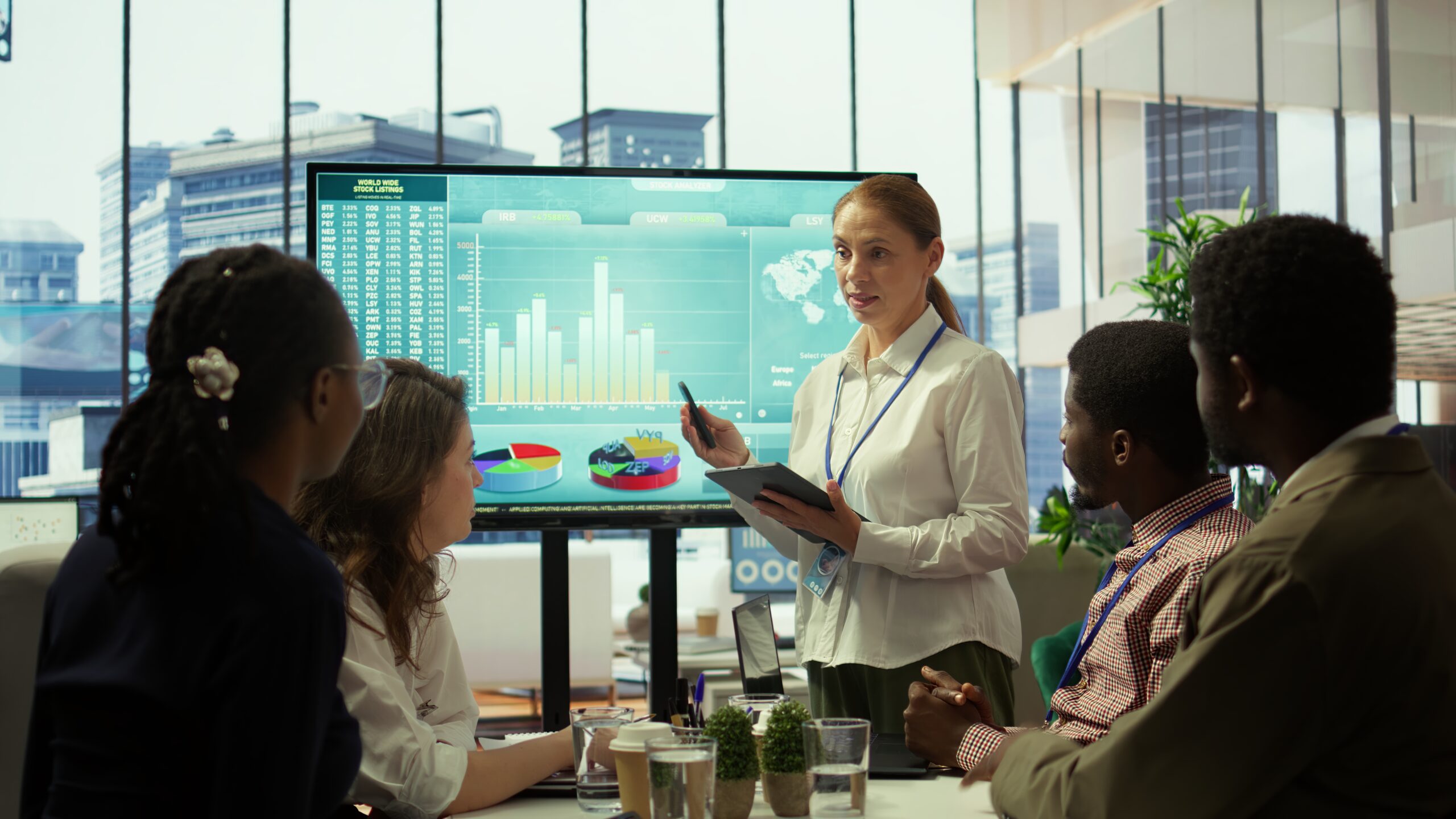 Em um escritório moderno com janelas amplas e vista para prédios urbanos, uma mulher de cabelo preso apresenta dados em um monitor grande para um grupo diverso de profissionais. O monitor exibe gráficos de barras, pizza e mapas com indicadores financeiros e estatísticos. Ela segura um tablet e uma caneta, enquanto explica as informações aos colegas sentados ao redor de uma mesa de reunião com notebooks, copos de água e pequenas plantas decorativas. O ambiente transmite colaboração, estratégia e análise de desempenho corporativo.