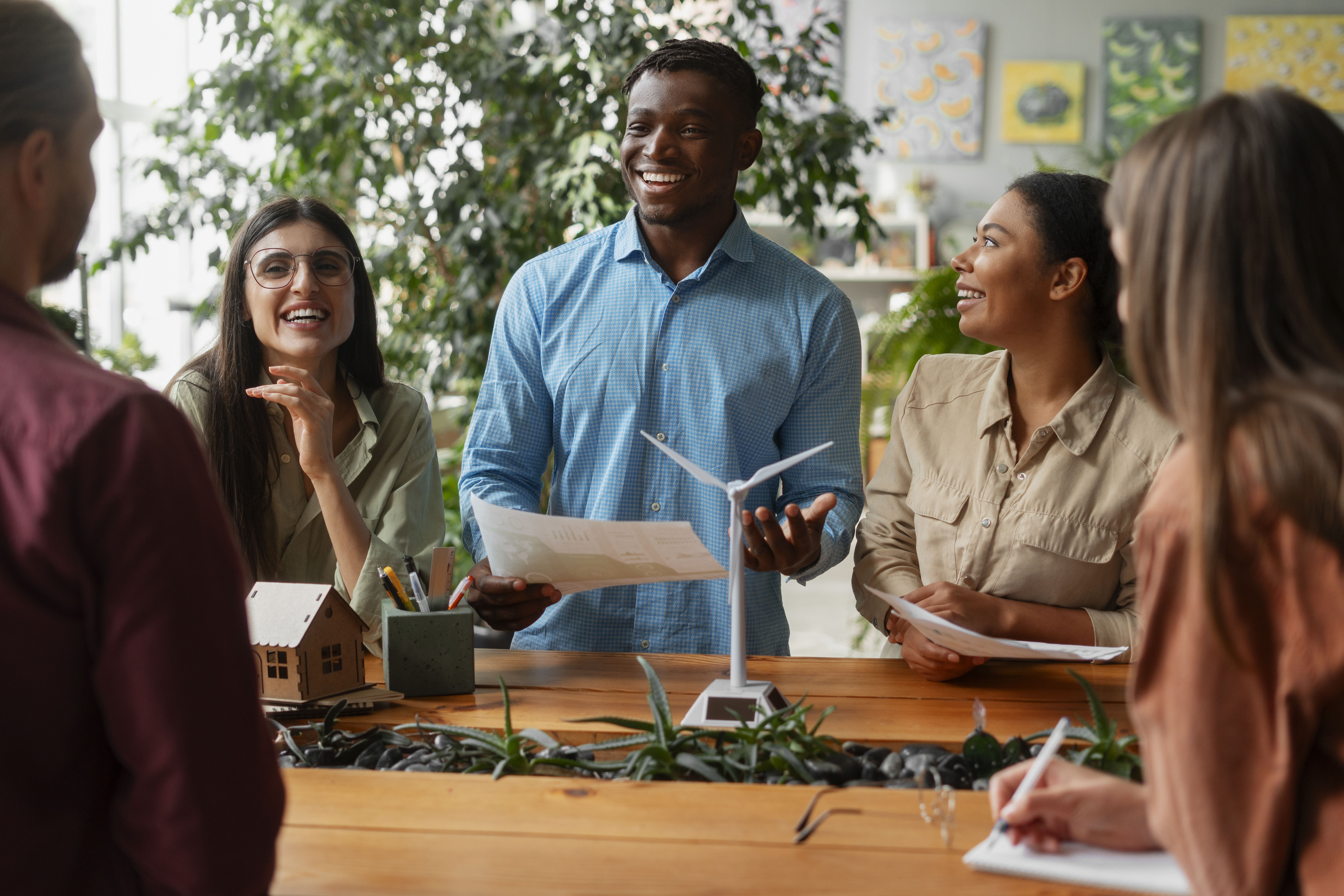 Grupo diverso de profissionais em reunião ao redor de uma mesa de madeira, sorrindo e trocando ideias sobre projetos de sustentabilidade. No centro da mesa há uma maquete de turbina eólica, plantas e materiais de escritório. Em destaque, um homem negro de camisa azul sorri enquanto segura uma folha de papel; ao seu lado, uma mulher negra de cabelo preso também sorri e participa da conversa. Há ainda uma mulher de óculos e cabelo longo e liso, usando camisa verde-clara, e outras duas pessoas parcialmente visíveis, contribuindo com anotações. O ambiente é claro, com vegetação ao fundo, transmitindo colaboração e engajamento em torno de soluções sustentáveis.
