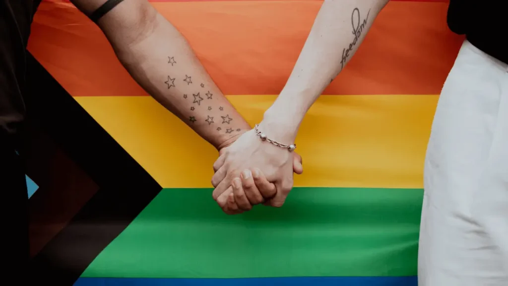 img lgbtfobia plurie 2 Duas mulheres de mãos dadas e atrás a bandeira do arco-íris